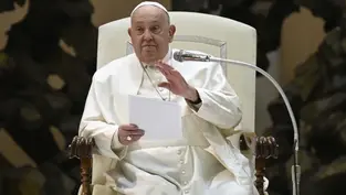 A man in white robes sits on a chair, holding papers and gesturing with his right hand. He is speaking in a formal setting with a microphone.