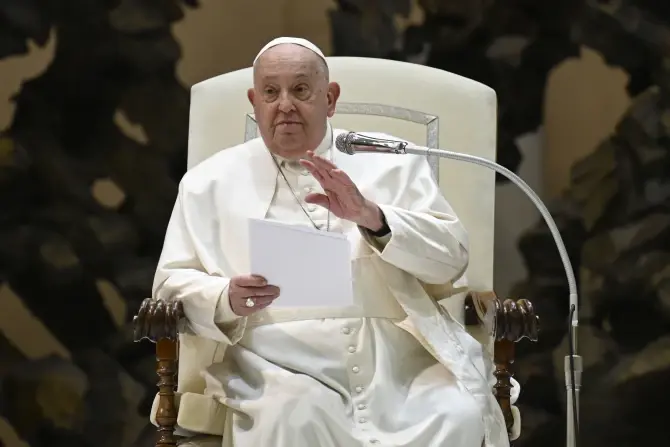 A man in white robes sits on a chair, holding papers and gesturing with his right hand. He is speaking in a formal setting with a microphone.