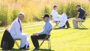 Two priests and men converse seated on chairs in a grassy field. One man wears a "NAVY" shirt. Sunny day with tall grass in the background.