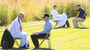 Two priests and men converse seated on chairs in a grassy field. One man wears a "NAVY" shirt. Sunny day with tall grass in the background.