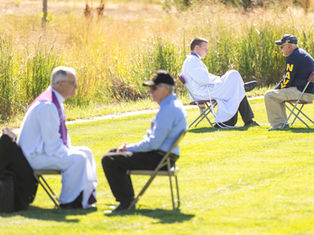 Two priests and men converse seated on chairs in a grassy field. One man wears a "NAVY" shirt. Sunny day with tall grass in the background.