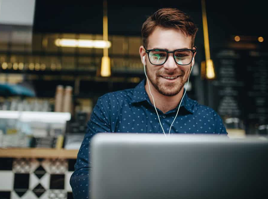 A Catholic man wearing glasses and headphones is focused on his laptop, reading the Denver Catholic.