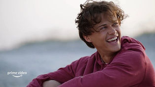 Young person in a red hoodie laughing on a beach with blurred background. Bright mood. "Prime Video" logo in the corner.