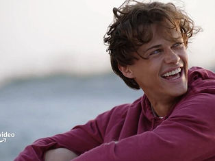 Young person in a red hoodie laughing on a beach with blurred background. Bright mood. "Prime Video" logo in the corner.