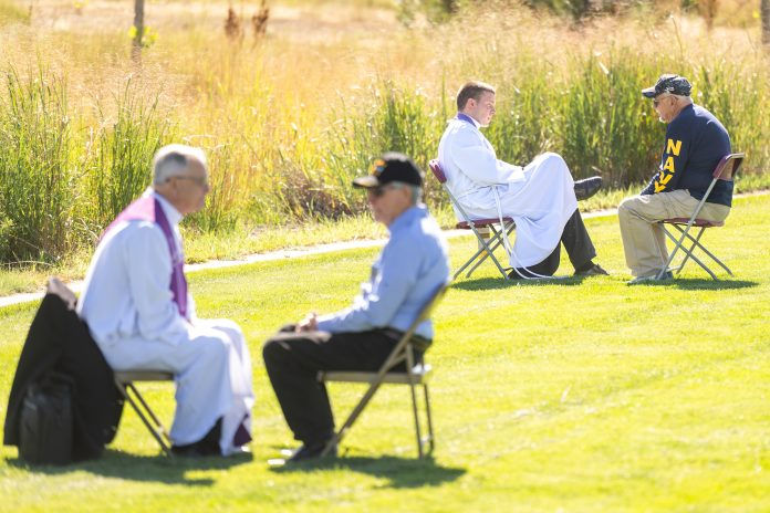 Two priests and men converse seated on chairs in a grassy field. One man wears a "NAVY" shirt. Sunny day with tall grass in the background.